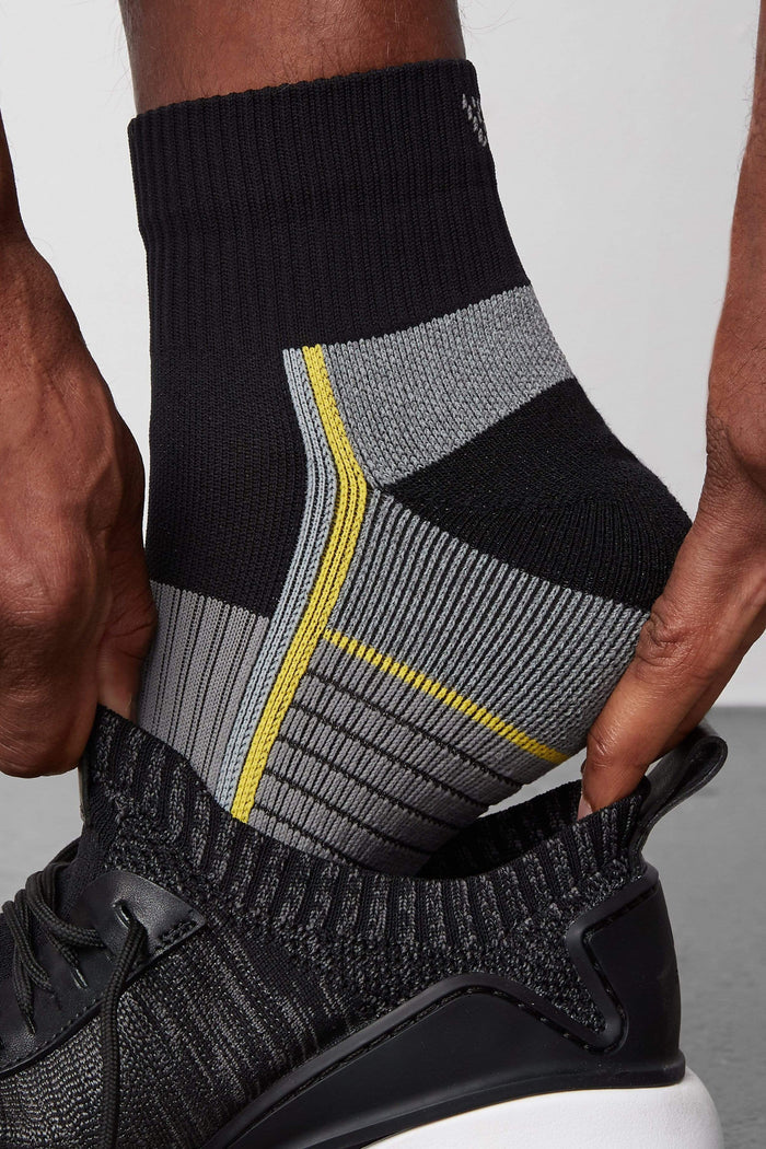 Side view of a man putting on a shoe and wearing AIRKNITx High Ankle Sock in True Black /Taxi