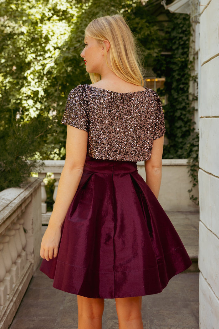 A woman with long blonde hair stands outside, wearing the Alexis Dress in Wine by Ivy City Co—its short sleeves and sequined, shiny burgundy fabric giving her the elegant look of a holiday party. She faces away, looking to the left, with greenery and white walls serving as the backdrop.
