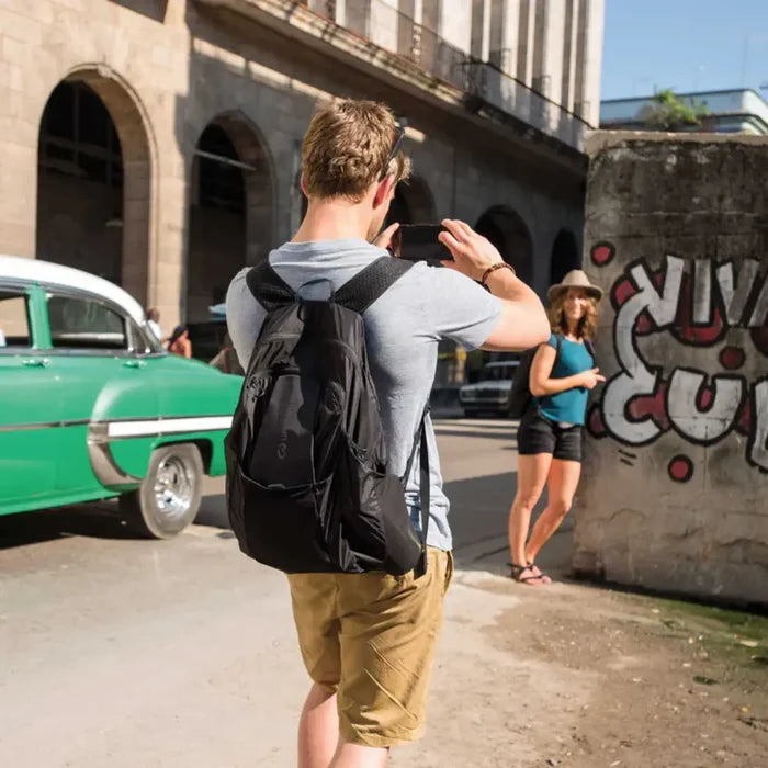Man with a Lifeventure Packable Backpack 25 Litres in black taking a photo of a woman near a vintage car and graffiti wall.