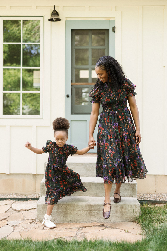 A woman and a young girl, both wearing matching Ivy City Co Mini Cordelia Dresses in Evergreen Vivid Meadow with puff sleeves and smocked bodices, hold hands and smile while stepping down from a porch in front of a house with a green door and white walls.