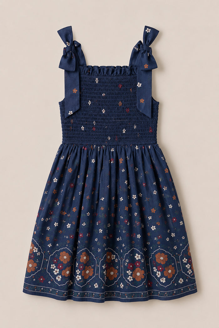 The Mini Rayna Dress in Midnight Cascading Floral by Ivy City Co is a navy blue, sleeveless chiffon dress featuring a smocked bodice and bow-tied shoulder straps. Its skirt showcases larger floral patterns and geometric designs along the hem, creating the perfect airy look for fall festivities.
