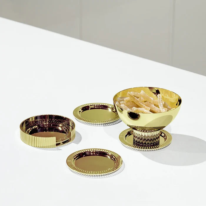 A shiny gold bowl filled with snacks rests on one of Anna New York’s Striations Coasters s/4 in Holder - Gold, accompanied by two additional empty Striations Coasters and a matching gold lid, all elegantly displayed on a white surface against a light background.