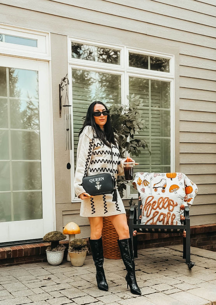 A woman stands on a front porch wearing a cream knit sweater dress with black geometric patterns, black knee-high boots, and carrying a black crossbody bag with 'QUEEN' on it. She is holding a coffee cup next to a decorated 'Hello Fall' chair.