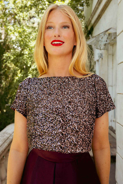 A woman with blonde hair and red lipstick stands outdoors, wearing the Ivy City Co Alexis Dress in Wine, which features short sleeves and a sparkling design with a dark skirt. Green foliage and a white building in the background enhance her elegant holiday party look.