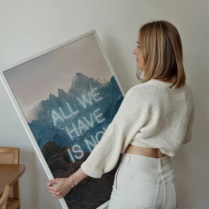 Woman holding a large framed print of misty mountains with glowing neon text reading All We Have Is Now.