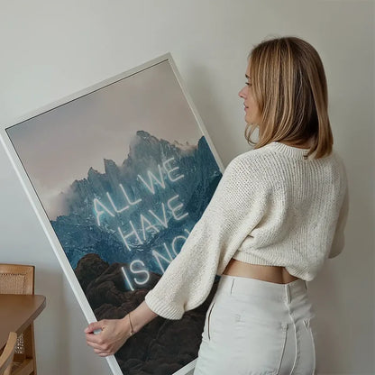 Woman holding a large framed print of misty mountains with glowing neon text reading All We Have Is Now.