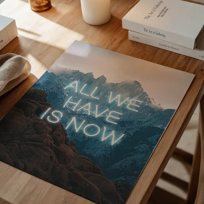 Glossy art print featuring glowing neon text All We Have Is Now over a dramatic mountain landscape on a wooden desk.