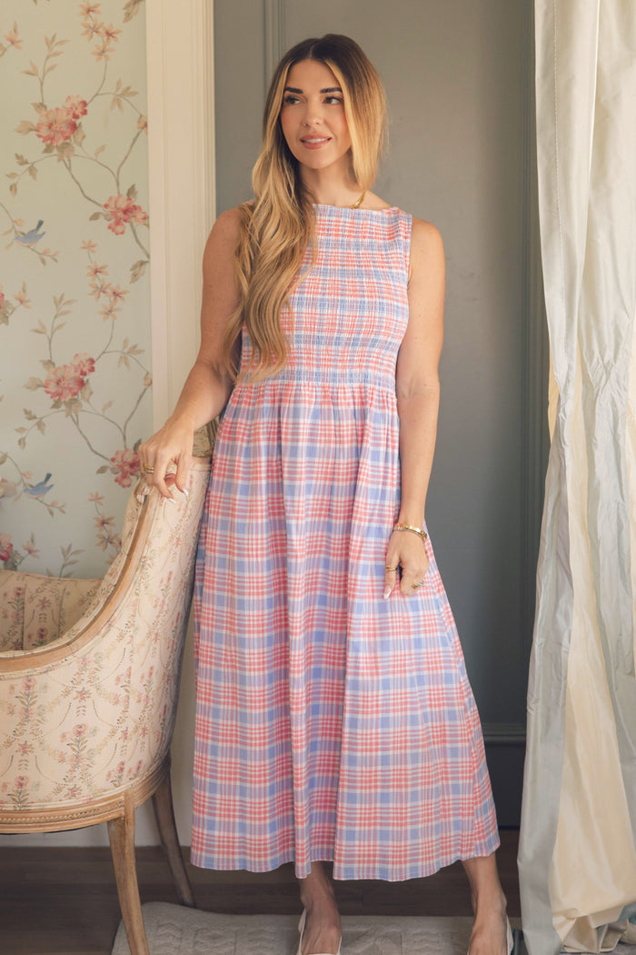 A woman with long light brown hair wears the Ivy City Co Ana Dress in Blossom Plaid, standing indoors by a floral chair. Floral wallpaper and soft curtains create a perfect spring backdrop for this charming plaid midi dress.