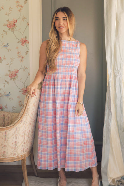 A woman with long light brown hair wears the Ivy City Co Ana Dress in Blossom Plaid, standing indoors by a floral chair. Floral wallpaper and soft curtains create a perfect spring backdrop for this charming plaid midi dress.