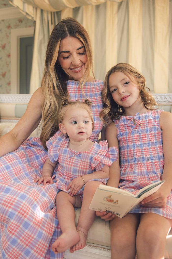 A woman and two young girls sit together on a couch, all wearing matching Ivy City Co Ana Dress in Blossom Plaid midi dresses. The older girl holds an open book while the woman smiles, their spring outfits catching the light.