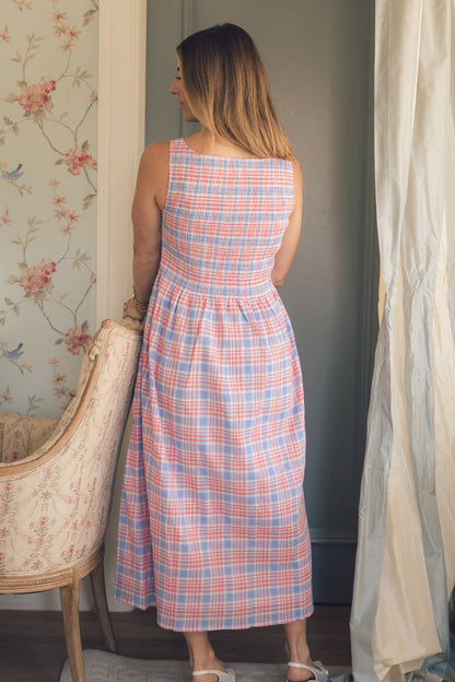 A woman with long hair, facing away, wears Ivy City Co’s Ana Dress in Blossom Plaid—a sleeveless, ankle-length, maternity-friendly dress in red, white, and blue. She stands by a floral armchair and a window with sheer curtains.