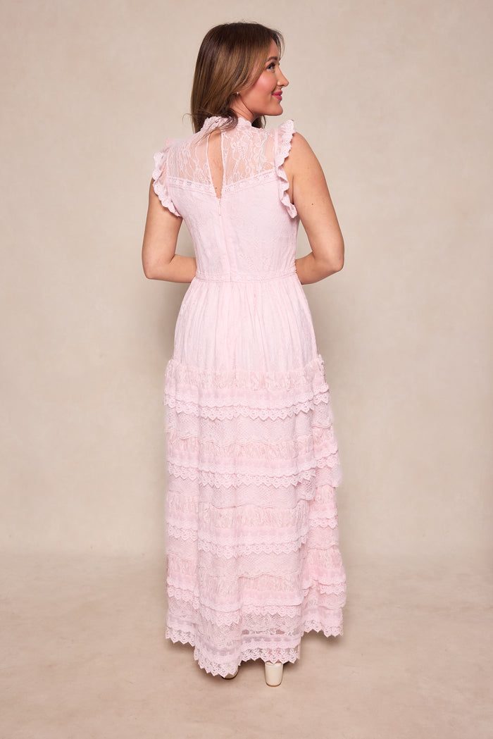 A woman faces away, wearing the Ivy City Co Annie Dress in Light Pink—a romantic silhouette with ruffled edges, tiered layers, a high neck, and sheer lace details—set against a neutral beige background.