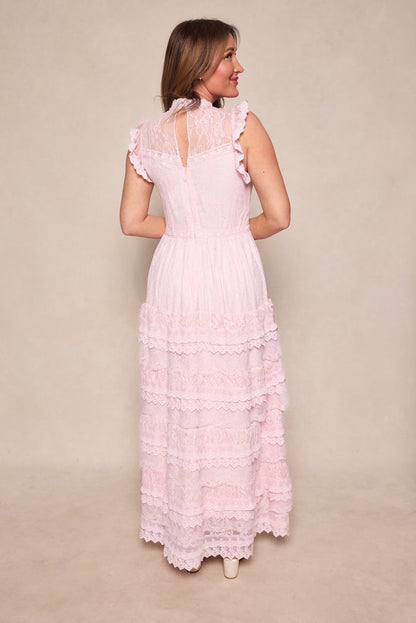 A woman faces away, wearing the Ivy City Co Annie Dress in Light Pink—a romantic silhouette with ruffled edges, tiered layers, a high neck, and sheer lace details—set against a neutral beige background.
