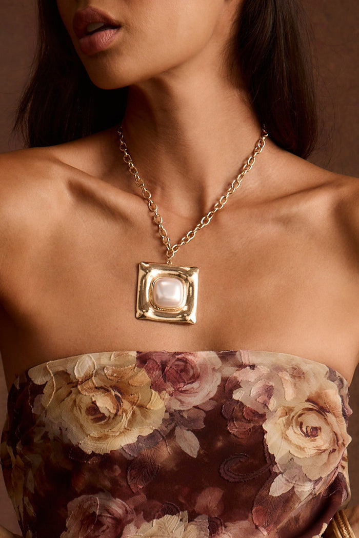 Gold chain necklace with a square pearl pendant worn over a strapless floral top.