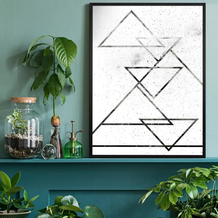 Framed abstract black triangle print on textured white paper, displayed on a teal shelf with plants.