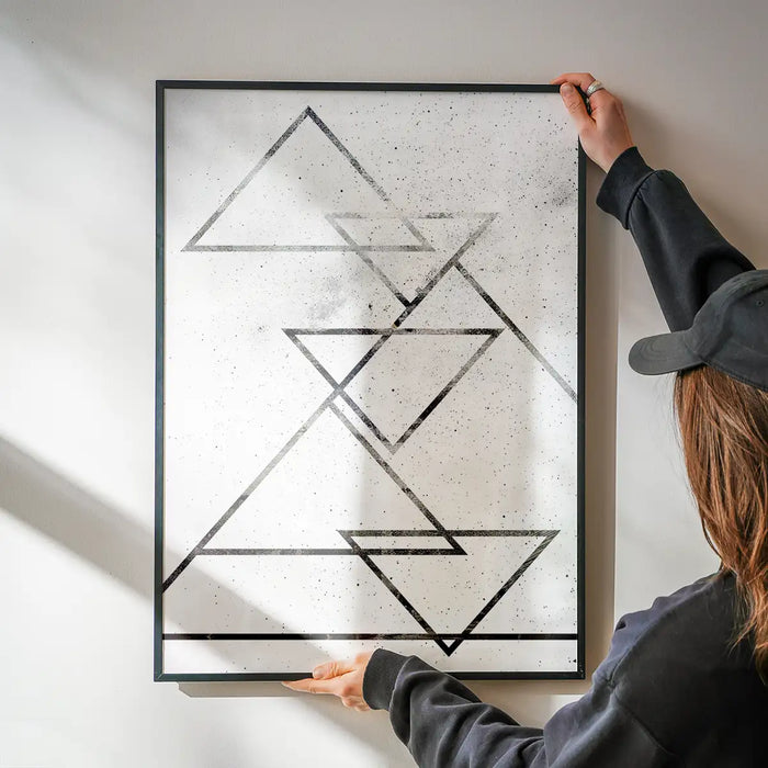 Abstract black triangle art print on textured white paper, held by hands against a wall.