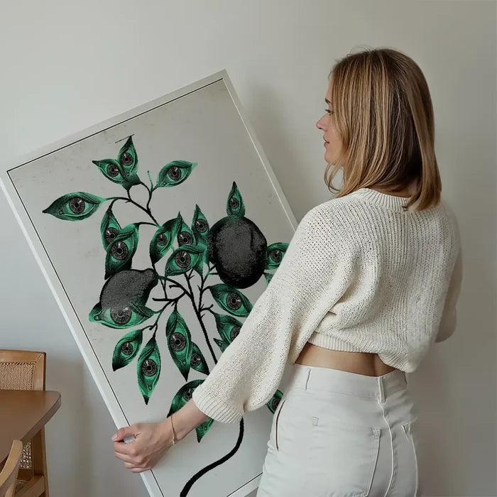 Woman holding a framed "Botanical Eyes" art print with dark fruit and green leaves featuring eyes.