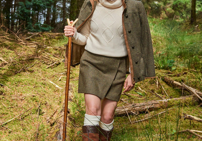 An image of a female model wearing the Dubarry women's Buckthorn Tweed Heath Skirt with concealed zip at the back and two functional welted front pockets. This is in a woodland setting and the model is holding a hiking stick.