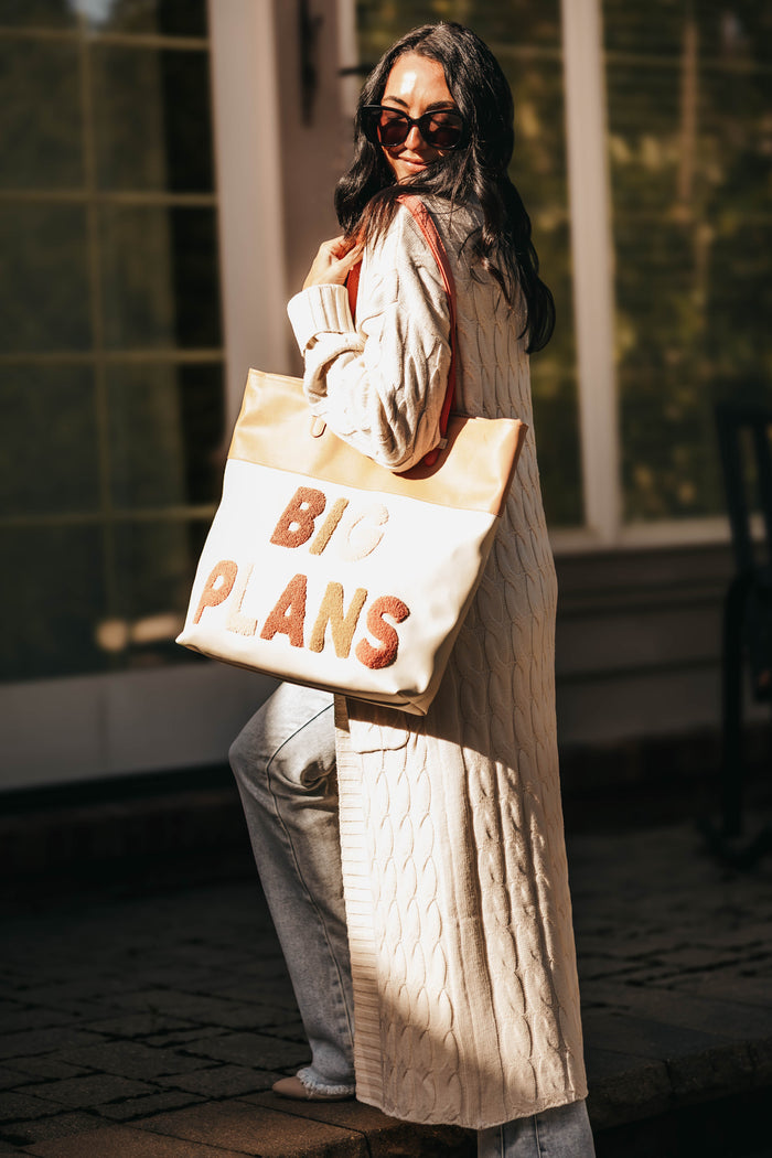 A woman in a cream cable-knit cardigan and sunglasses carries a large cream canvas tote bag with 'BIG PLANS' written on it in orange-brown letters, set against a warm, sunlit outdoor background.