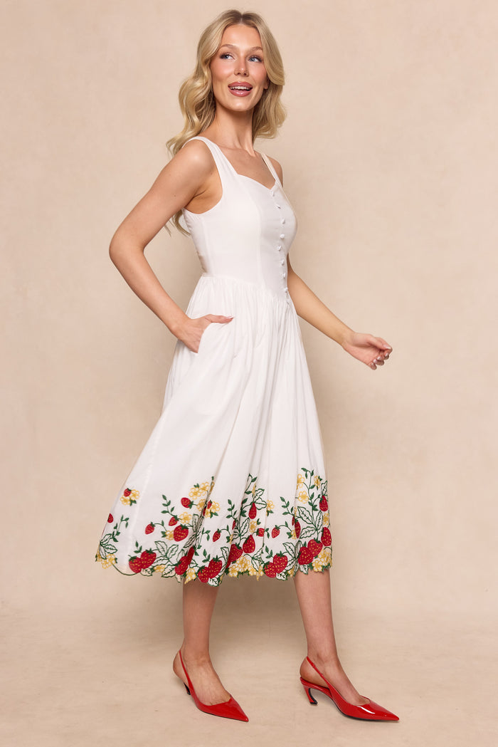 A smiling woman with long blonde hair wears the Ivy City Co Clarissa Dress in Strawberry, featuring sleeveless white cotton and colorful floral embroidery along the hem, paired with red pointed heels against a light beige background.
