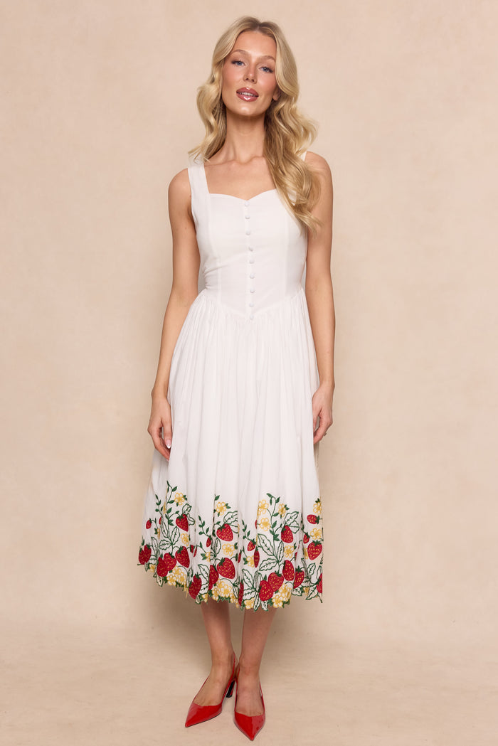 A woman wears the Ivy City Co Clarissa Dress in Strawberry—a sleeveless white cotton graduation dress with front buttons and colorful floral embroidery at the hem—paired with red pointed-toe heels, standing before a neutral background.