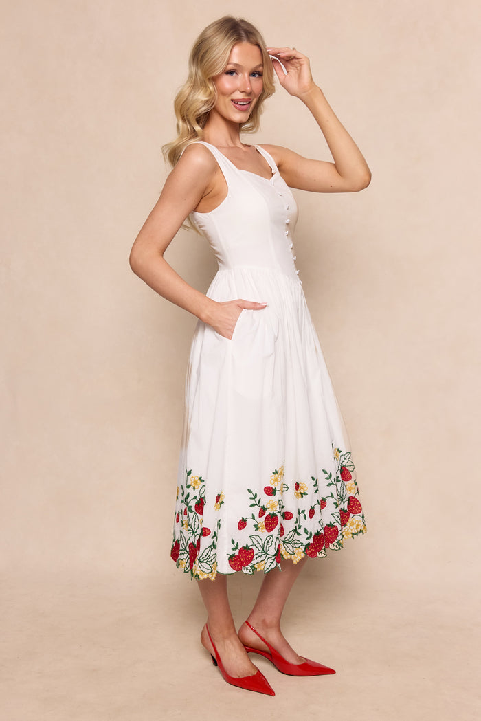 A woman smiles against a beige background, wearing the Ivy City Co Clarissa Dress in Strawberry, a sleeveless cotton dress with colorful floral embroidery along the hem, paired with red pointed-toe heels. Her long blonde hair is styled in waves.
