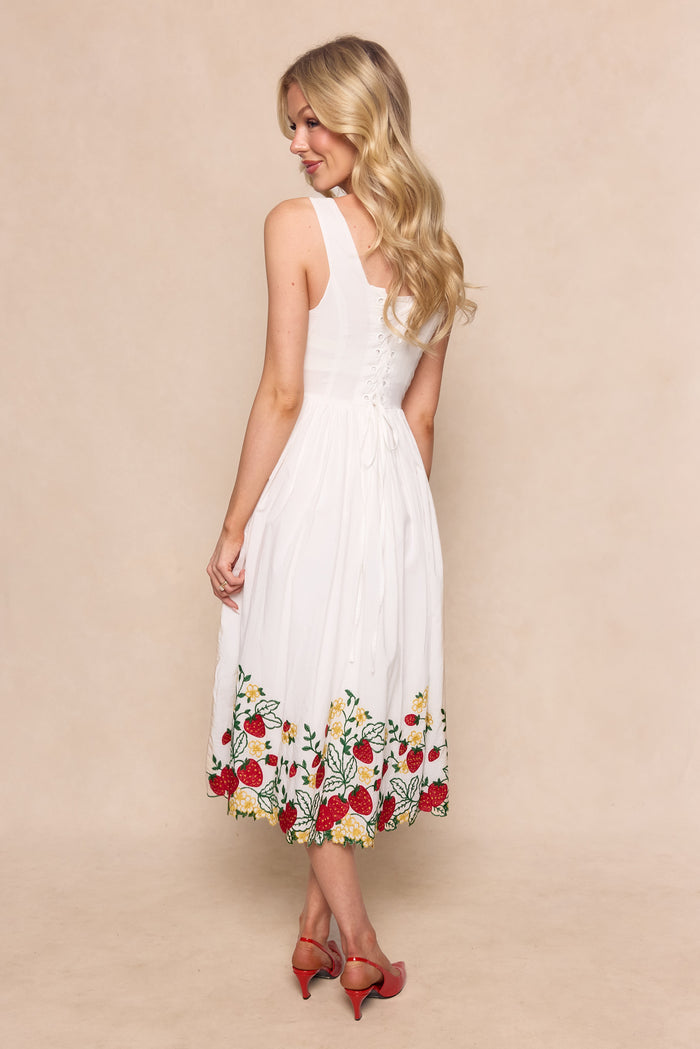 A woman with long blonde hair stands facing away, wearing the Ivy City Co Clarissa Dress in Strawberry, a cotton graduation dress with a lace-up corset back and colorful floral embroidery at the hem, paired with red heeled sandals against a light beige background.