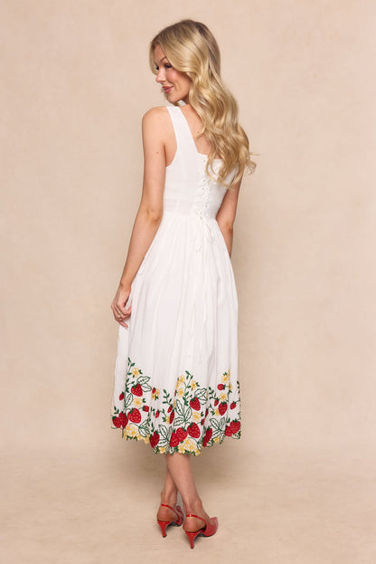 A woman with long blonde hair stands facing away, wearing the Ivy City Co Clarissa Dress in Strawberry, a cotton graduation dress with a lace-up corset back and colorful floral embroidery at the hem, paired with red heeled sandals against a light beige background.
