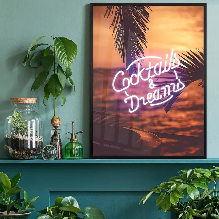 Cocktails and Dreams neon sign art print featuring palm trees at sunset, displayed in a black frame on a teal shelf.