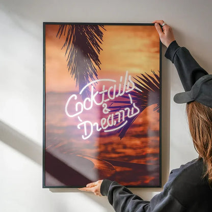 Cocktails and Dreams neon sign art print featuring palm trees at sunset in a black frame held against a white wall.