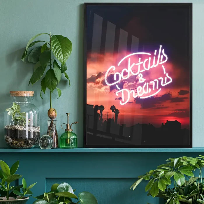 Framed neon Cocktails and Dreams print featuring a vibrant sunset silhouette, displayed on a teal shelf with indoor plants.