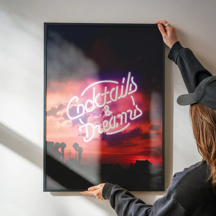 Glossy framed print featuring a glowing Cocktails and Dreams neon sign over a vibrant sunset with palm tree silhouettes.