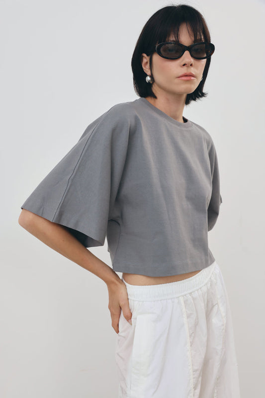 a gray cropped t-shirt with wide, voluminous sleeves and white pants, styled with black sunglasses against a plain light gray background.