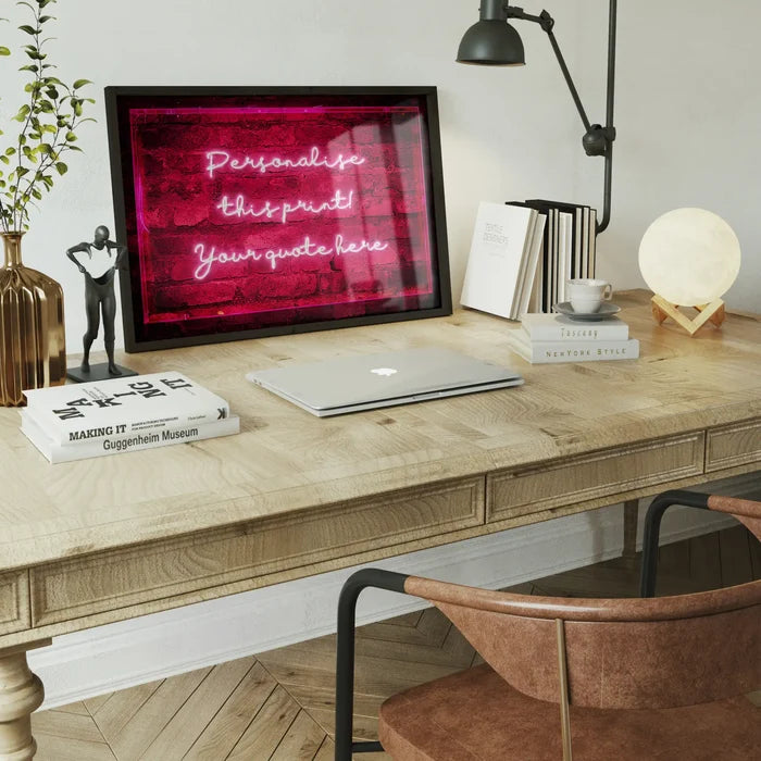 Framed neon-style print featuring glowing pink cursive text on a red brick background, displayed on a wooden desk.