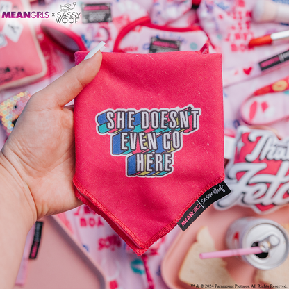 A hand holds the Sassy Woof Dog Bandana - Mean Girls™ She Doesn't Even Go Here, featuring bold, colorful lettering. The background is filled with playful pink dog accessories, cups, snacks, and more.