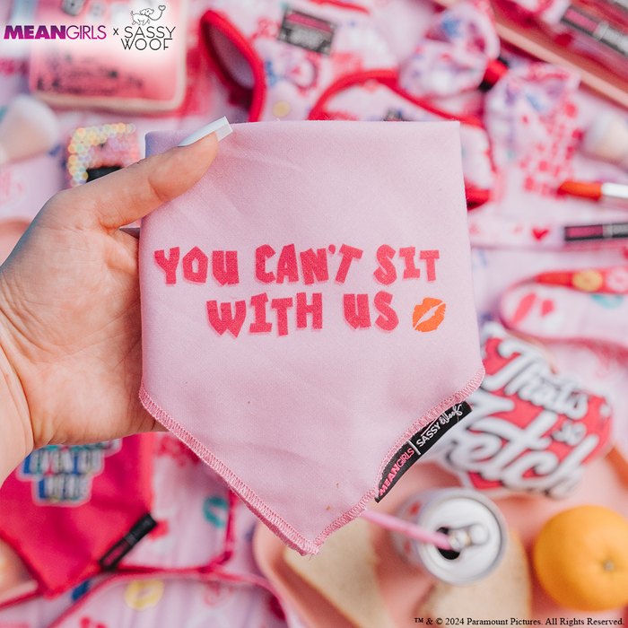 A hand holds the Sassy Woof Dog Bandana - Mean Girls™ You Can't Sit With Us, featuring bold text and a lip print. In the background are stylish dog accessories and pink treats inspired by the iconic film.