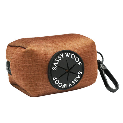 Brown rectangular dog waste bag holder with a black circular “Sassy Woof” label, featuring a plastic clip for easy attachment to your leash or bag. This accessory is part of the Sassy Woof Dog Collar Three Piece Bundle - Teddy from the adorable teddy collection.