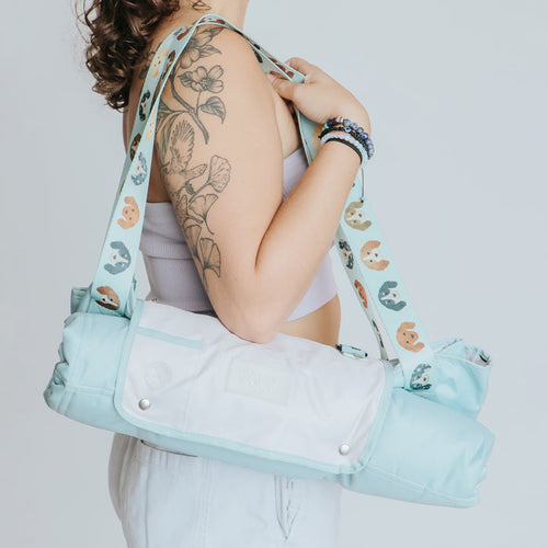 A person with a floral tattoo carries a light blue and white yoga mat bag over their shoulder, wearing a lavender top and white pants, while the Sassy Woof Dog Picnic Blanket - Dainty Dachshunds is tucked under their arm for an outdoor session.