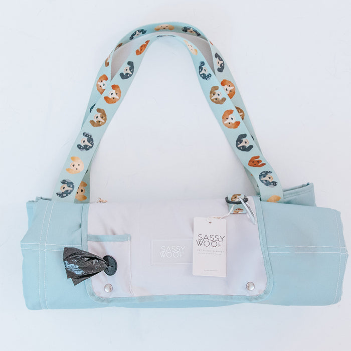 A Sassy Woof light blue and white bag with a colorful dog print strap, front waste bag dispenser, and tag—ideal for carrying your Dog Picnic Blanket - Dainty Dachshunds by Sassy Woof on any outing. Shown on a white background.