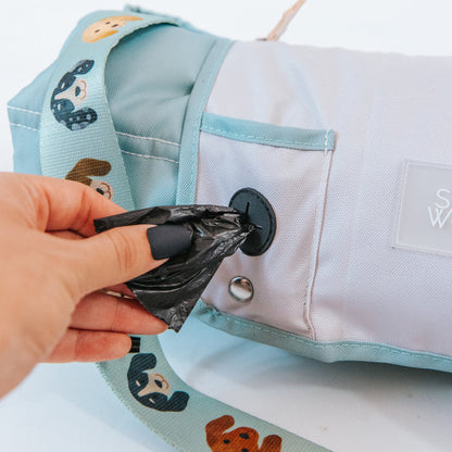 A hand pulls a black waste bag from the dispenser pocket on the Sassy Woof Dog Picnic Blanket - Dainty Dachshunds, a light blue and white outdoor dog blanket bag with a cartoon dachshund strap.