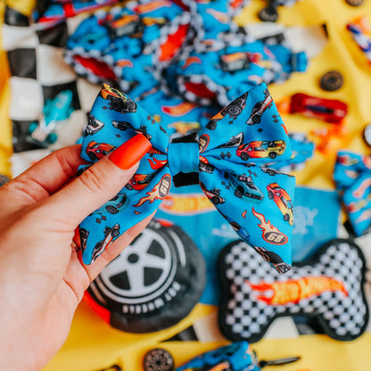 A hand with bright orange nail polish holds the Sassy Woof Dog Sailor Bow - Hot Wheels™, surrounded by matching Hot Wheels™ dog accessories like an adjustable harness and collar, all arranged on a yellow surface.