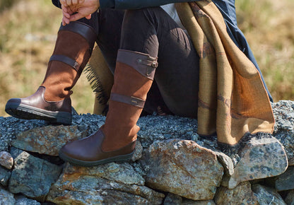 An outdoor on model image of A pair of Dubarry Dunmore Country boots, walnut brown coloured leathered knee high boots with adjustable buckle, with the model sitting on a stone wall.