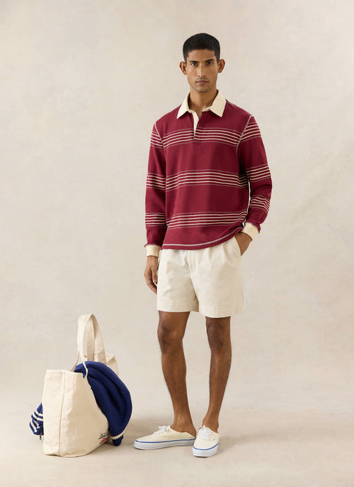 Percival Editor burgundy cotton rugby polo with cream stripes and stab stitching, styled with cream shorts and a tote bag.