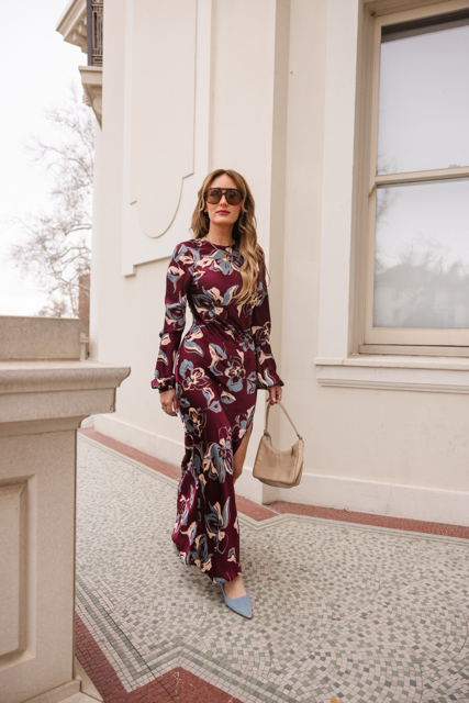 A woman wearing the Ivy City Co Elyse Dress in Burgundy Floral, sunglasses, and light blue shoes walks outside by a cream-colored building, carrying a beige handbag.