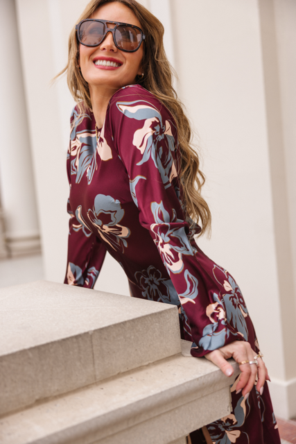Smiling broadly, a woman with long wavy hair and sunglasses leans on a stone railing against a light architectural backdrop, wearing the Elyse Dress in Burgundy Floral by Ivy City Co.