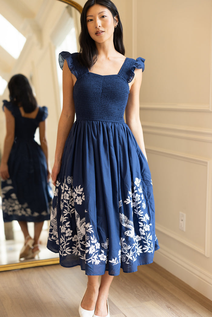 A woman stands indoors wearing the Ivy City Co Finch Dress in Blue, a sleeveless, knee-length cotton dress with white embroidery. Her reflection appears in a large mirror. She pairs the dress with white shoes and has long dark hair.
