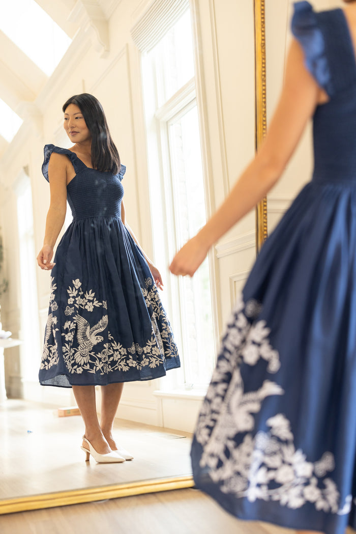 A woman in the Finch Dress in Blue by Ivy City Co admires herself in a large mirror. She stands on light wood flooring in a bright, sunlit room with tall windows, pairing the dress with white heels.