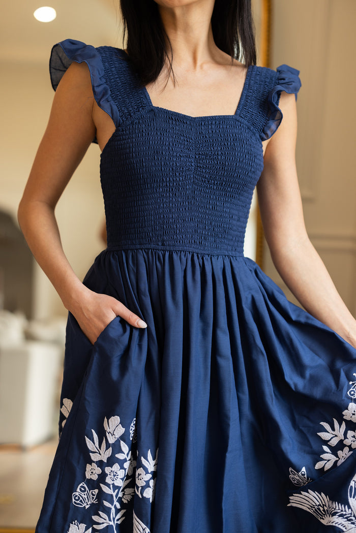 A woman stands indoors in the Finch Dress in Blue by Ivy City Co, a deep blue cotton dress with ruffled straps, white embroidery near the hem, and handy pockets, holding her skirt and slipping one hand into a pocket.