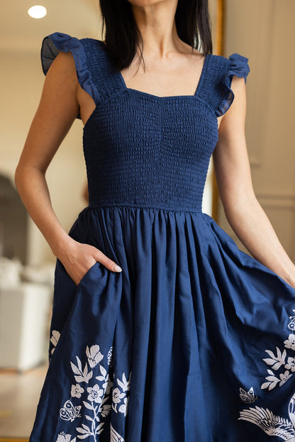 A woman stands indoors in the Finch Dress in Blue by Ivy City Co, a deep blue cotton dress with ruffled straps, white embroidery near the hem, and handy pockets, holding her skirt and slipping one hand into a pocket.