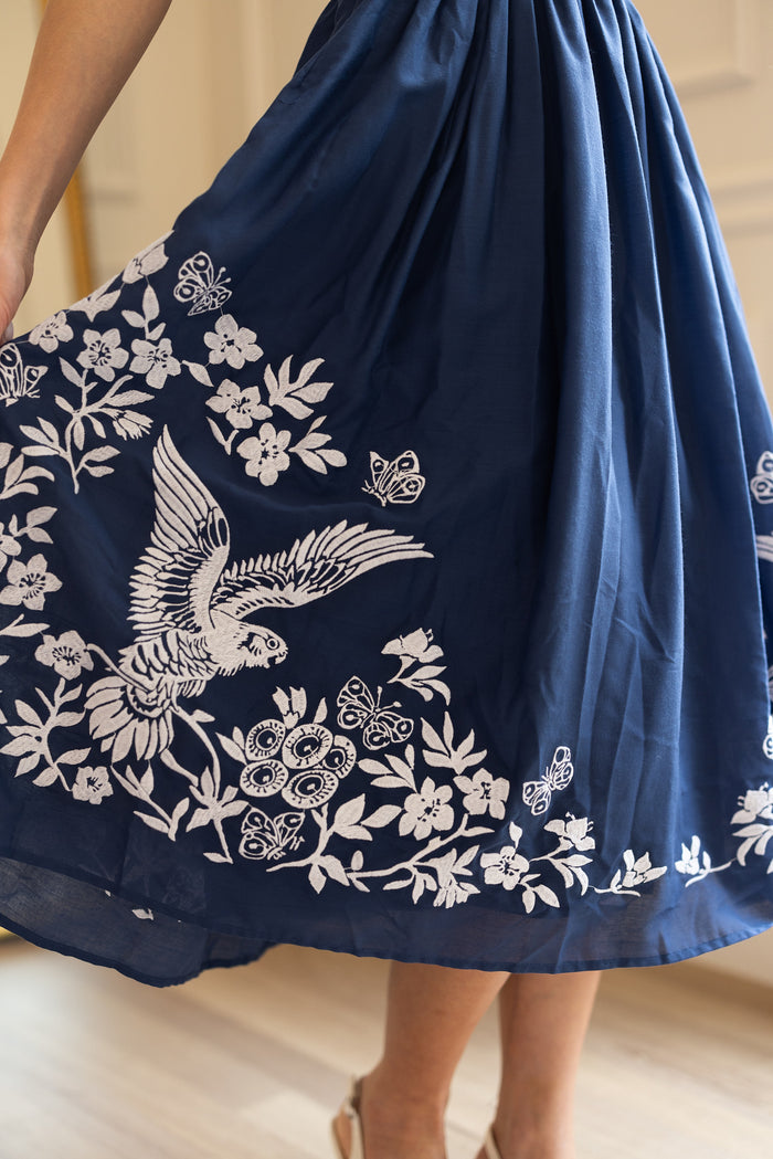 A person indoors wears the Finch Dress in Blue by Ivy City Co, featuring deep blue fabric with white embroidery of an owl, flowers, leaves, and butterflies.